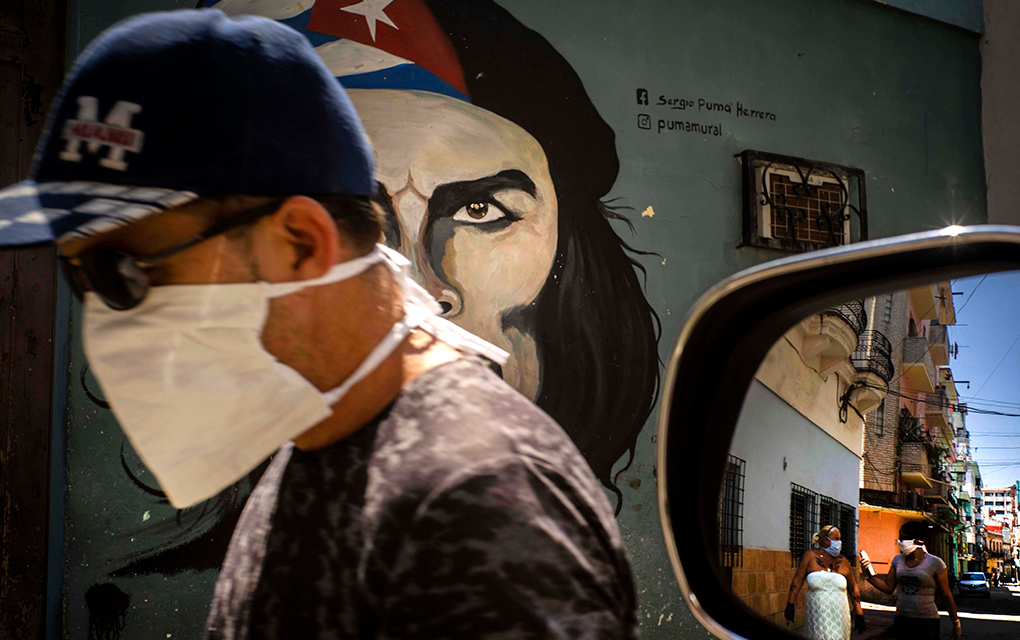 Un hombre con una mascarilla camina frente a un mural de Ernesto Che Guevara, mientras que otros peatones aparecen reflejados en el espejo de un vehículo en La Habana, Cuba / Foto: AP