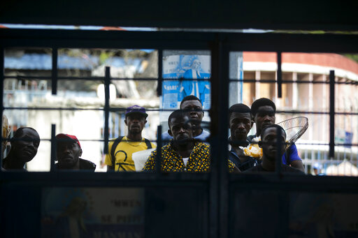 Varios individuos que no lucen tapabocas observan a través de un portón un partido entre Haití y Belice por las eliminatorias de la Copa Mundial de fútbol en Puerto Príncipe el 25 de marzo del 2021. ( AP Photo/ Dieu Nalio Chery)
